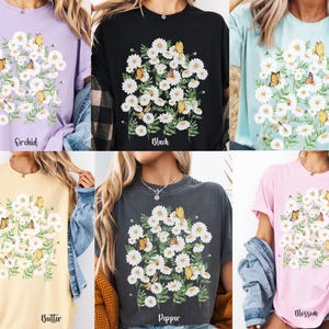 以下が含まれることがあります： さまざまな色のTシャツ。それぞれに白いデイジーと蝶のフローラルデザインが施されています。シャツの色は、オーキッド、ブラック、シャンブレー、バター、ペッパー、ブロッサムです。