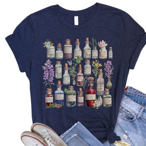 May include: A navy blue t-shirt with a graphic print of various glass bottles filled with colorful liquids and herbs. The bottles are arranged in a circular pattern and have labels with handwritten text.