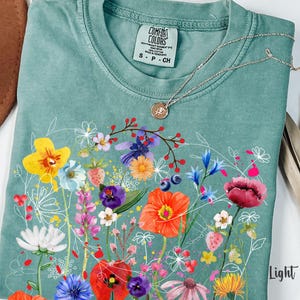 May include: A light green t-shirt with a floral print featuring a variety of colorful flowers, including red, pink, yellow, and blue. The shirt has a crew neck and short sleeves. The text "COMFORT COLORS" is visible on the shirt.