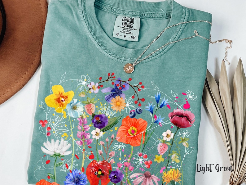 May include: A light green t-shirt with a floral print featuring a variety of colorful flowers, including red, pink, yellow, and blue. The shirt has a crew neck and short sleeves. The text "COMFORT COLORS" is visible on the shirt.