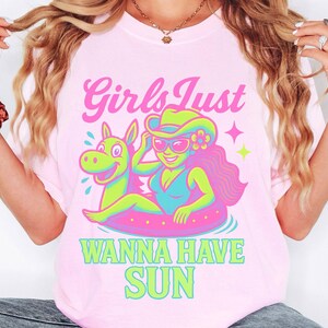 May include: Pink t-shirt with a graphic of a woman in a swimsuit and cowboy hat floating on a pool float with a cartoon horse. The text "Girls Just Wanna Have Sun" is above the graphic in pink and green.