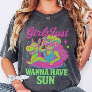 May include: Dark gray t-shirt with a colorful graphic. The graphic features a cartoon woman in a swimsuit and cowboy hat, floating on a pool float with a horse. Text reads "Girls Just Wanna Have Sun."