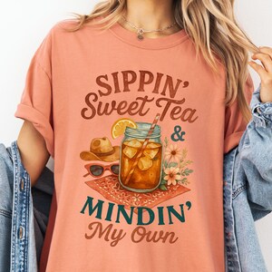 Może przedstawiać: Koszulka w kolorze brzoskwiniowym z napisem "Sippin' Sweet Tea & Mindin' My Own" w brązowych, zielonych i białych literach. Projekt przedstawia szklankę lodowatej herbaty z plasterkiem cytryny, słomką i kwiatami.