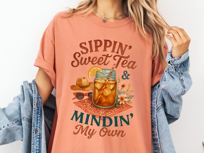 Może przedstawiać: Koszulka w kolorze brzoskwiniowym z napisem "Sippin' Sweet Tea & Mindin' My Own" w brązowych, zielonych i białych literach. Projekt przedstawia szklankę lodowatej herbaty z plasterkiem cytryny, słomką i kwiatami.