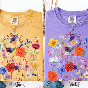 May include: Two t-shirts with a floral design. The shirt on the left is mustard yellow with a white floral design. The shirt on the right is violet with a white floral design. Both shirts have the text "Comfort Colors" on the tag.