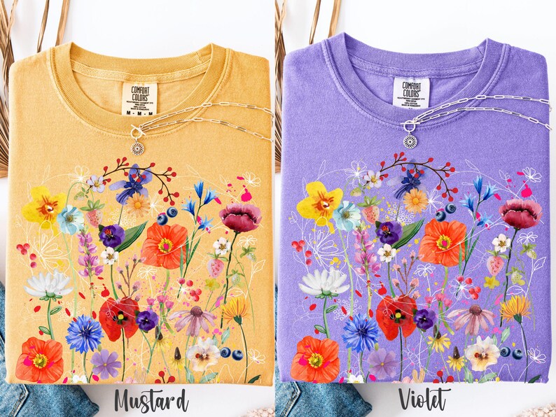 May include: Two t-shirts with a floral design. The shirt on the left is mustard yellow with a white floral design. The shirt on the right is violet with a white floral design. Both shirts have the text "Comfort Colors" on the tag.