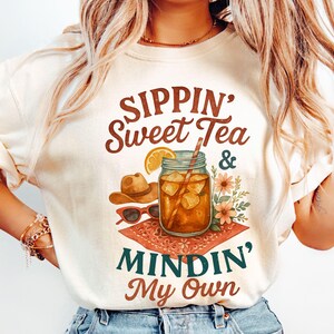 Może przedstawiać: Biały T-shirt z napisem "Sippin' Sweet Tea & Mindin' My Own" w brązowych i zielonych literach. Projekt zawiera r&oacute;wnież szklankę lodowatej herbaty z plasterkiem cytryny, słomką i kowbojskim kapeluszem.
