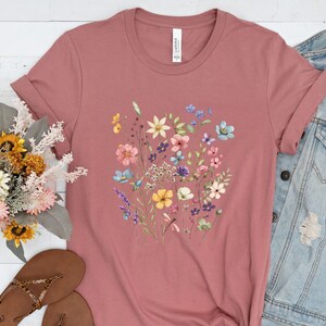 May include: A mauve t-shirt with a floral print featuring a variety of colorful flowers, including pink, blue, yellow, and purple.