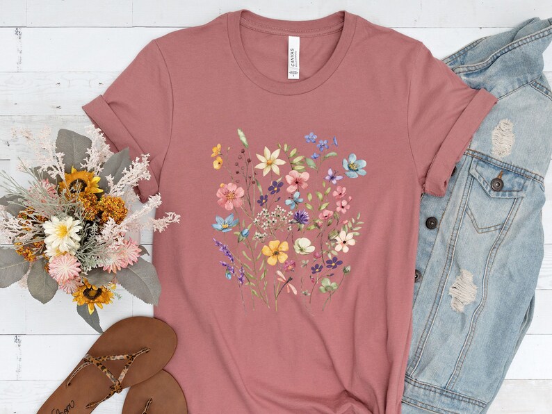 May include: A mauve t-shirt with a floral print featuring a variety of colorful flowers, including pink, blue, yellow, and purple.