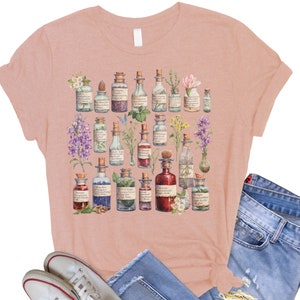 May include: A pink t-shirt with a floral design featuring a variety of glass bottles filled with colorful liquids. The bottles are surrounded by flowers and leaves. The design is reminiscent of a vintage apothecary.