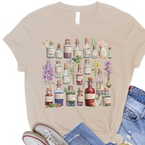 May include: A beige t-shirt with a vintage illustration of various glass bottles filled with colorful liquids and surrounded by flowers and herbs.