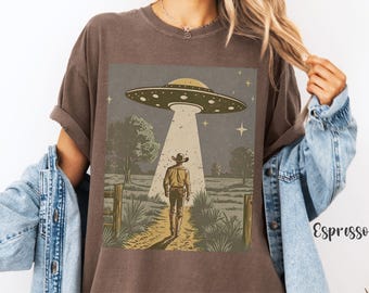 May include: A brown t-shirt with a vintage-style illustration of a cowboy standing in a field with a UFO hovering above him. The UFO is emitting a beam of light. The text on the shirt is "Espresso".
