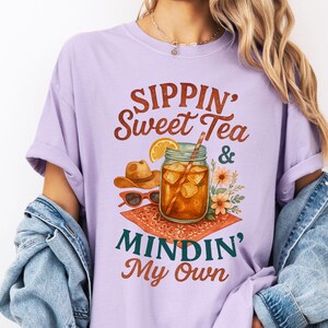 Może przedstawiać: Fioletowy t-shirt z napisem "Sippin' Sweet Tea & Mindin' My Own" w brązowych literach. Projekt zawiera szklankę lodowatej herbaty z plasterkiem cytryny i słomką, kowbojski kapelusz i okulary przeciwsłoneczne.