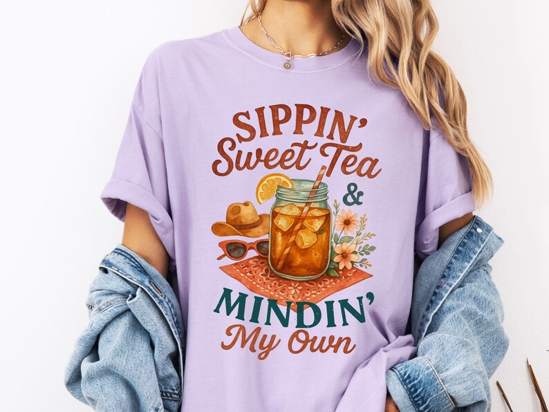 Może przedstawiać: Fioletowy t-shirt z napisem "Sippin' Sweet Tea & Mindin' My Own" w brązowych literach. Projekt zawiera szklankę lodowatej herbaty z plasterkiem cytryny i słomką, kowbojski kapelusz i okulary przeciwsłoneczne.
