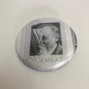 May include: A black and white button with a photo of Charles Bukowski holding a book. The button has the text "HORSE MEAT" printed on it.