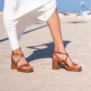 Può includere: Sandali con plateau in pelle marrone con cinturini incrociati e tacco in legno spesso. I sandali hanno una suola platform e un cinturino alla caviglia con chiusura a fibbia. Indossati con un vestito bianco.