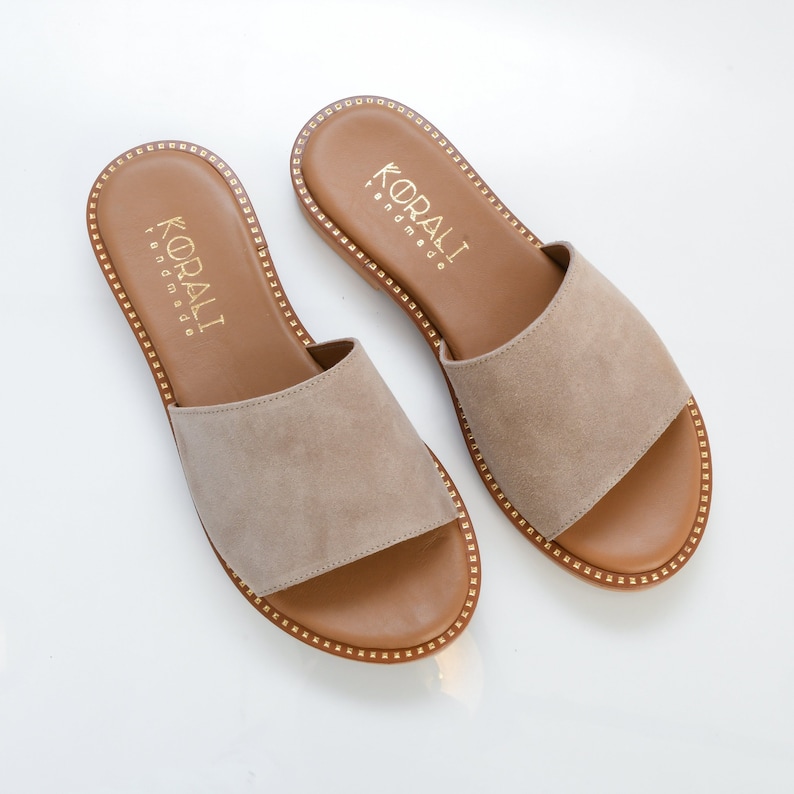 May include: A pair of beige suede sandals with a brown leather sole and gold trim. The sandals have a simple design and are perfect for summer.