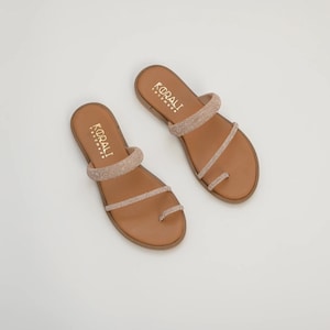May include: A pair of brown sandals with a toe ring and two straps adorned with sparkling embellishments. The sandals have a flat sole and the brand name "KORALI" is printed on the footbed.