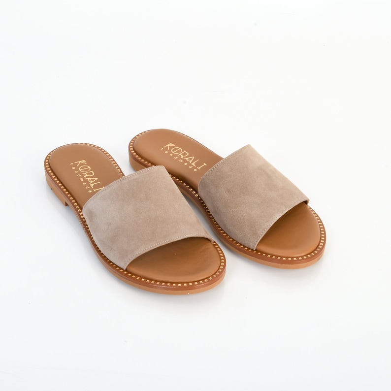 May include: A pair of beige suede slide sandals with a brown leather sole and gold stitching. The sandals have the brand name "KORALI" printed on the straps.