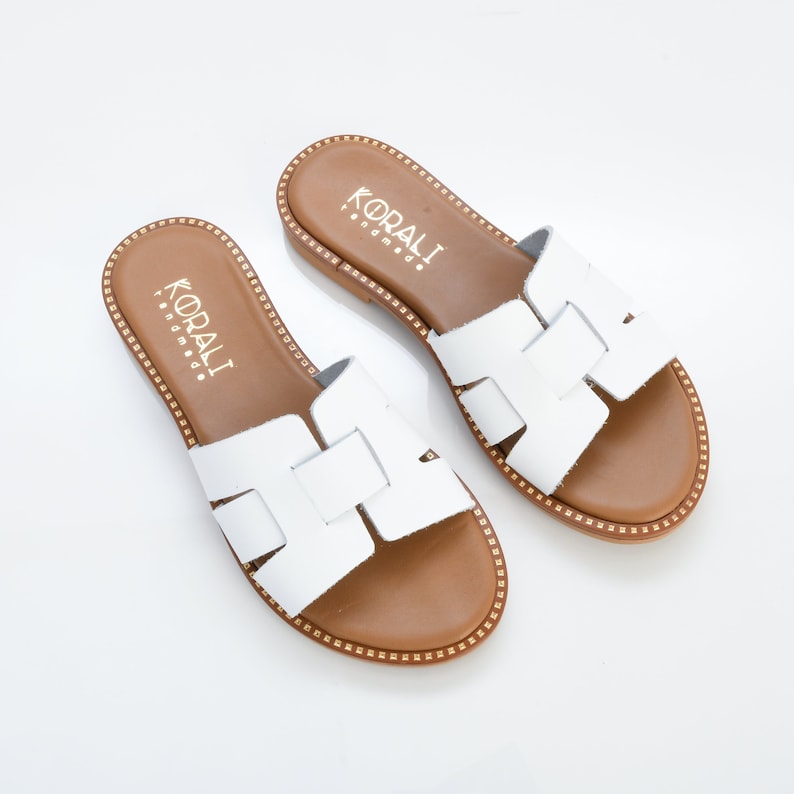Pu&ograve; includere: Sandali in pelle bianca con suola marrone e dettagli dorati. I sandali hanno un design a cinturino e il nome del marchio "KORALI" &egrave; stampato sulla soletta.