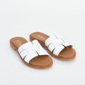 Pu&ograve; includere: Sandali in pelle bianca con suola in pelle marrone e design a taglio. I sandali hanno il nome del marchio "KORALI" stampato sui cinturini.