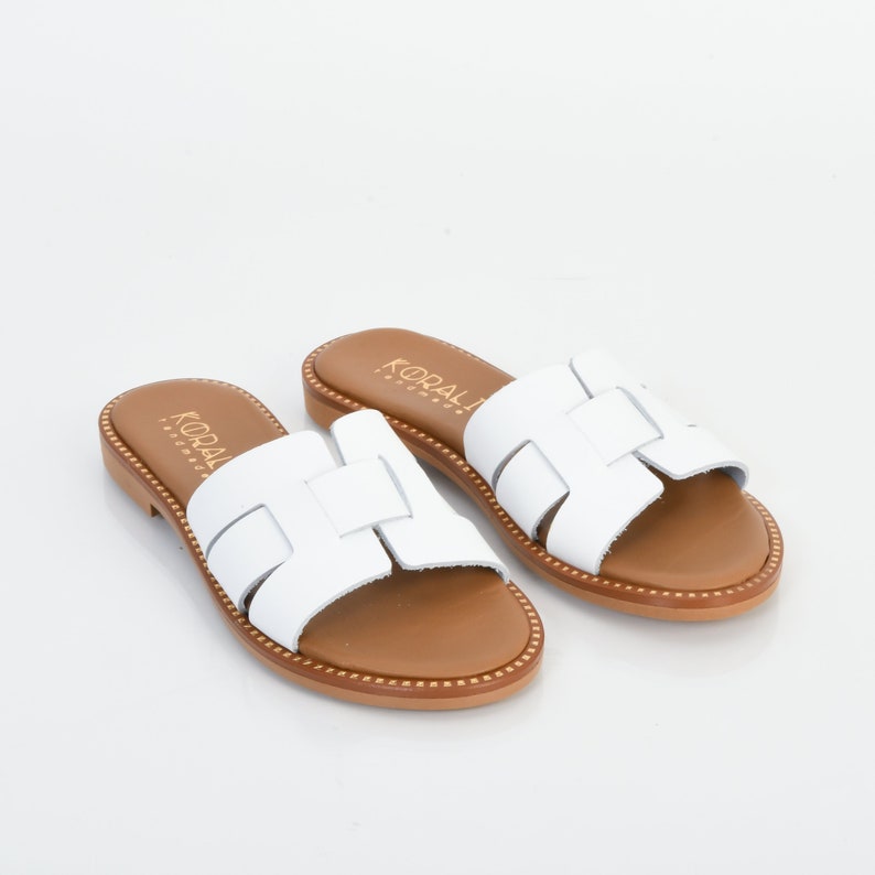 Pu&ograve; includere: Sandali in pelle bianca con suola in pelle marrone e design a taglio. I sandali hanno il nome del marchio "KORALI" stampato sui cinturini.