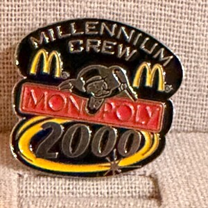 May include: A collectible pin featuring the text "Millennium Crew" and "Monopoly 2000". The pin has a black background with yellow and red accents, including the McDonald's logo. The Monopoly man is also featured.