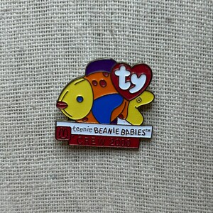 May include: A colorful enamel pin featuring a fish design with yellow, orange, blue, and purple accents. The pin includes the text "ty" in a heart shape and "teenie BEANIE BABIES" and "CREW 2000".