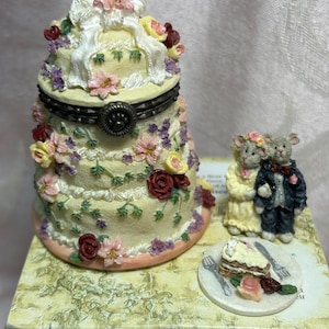May include: A decorative, tiered cake-shaped trinket box with floral embellishments and a bow on top. A pair of mouse figurines in wedding attire stand beside a plate with a slice of cake. The box is on a decorative box.
