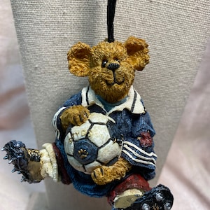 May include: A brown teddy bear wearing a blue and white soccer jersey and black cleats, holding a white soccer ball with black markings. The ball has the word "PLAY" printed on it.