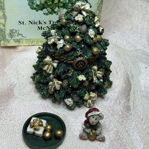 以下が含まれることがあります： ヒンジ付きの蓋が付いた装飾的なクリスマスツリーの置物で、金色のオーナメント、白いリボン、松ぼっくりで飾られています。その横には、白いギフトボックスと金色の球体が乗った小さな丸い緑色のトレイと、赤い帽子をかぶったネズミの置物が置かれています。