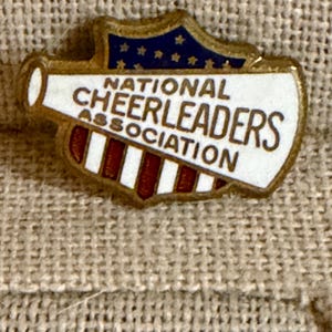 May include: A vintage pin with the words "NATIONAL CHEERLEADERS ASSOCIATION" in white lettering. The pin has a gold border, a blue top with gold stars, and a red and white striped bottom. The pin is shaped like a megaphone.