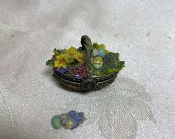 May include: A small, decorative basket filled with colorful flowers, fruit, and eggs. The basket is made of woven material and has a brown handle. There is a small, green and purple figurine of a bird or animal lying next to the basket.