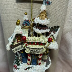 Puede incluir: Un adorno navideño miniatura blanco, cubierto de nieve, que representa un hospital del Polo Norte con un letrero que dice "Santa's Re-Patch North Pole Hospital". El adorno tiene un diseño de bastón de caramelo rojo y blanco y una puerta azul.