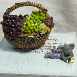 以下が含まれることがあります： 緑と紫のブドウの房で満たされた編みかご型の小物入れで、足跡の刻印がある金属製の留め具が付いています。その横には、紫色のズボンを履いてブドウの房を持っている小さな灰色のネズミのフィギュアが置かれています。