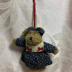 Pode incluir: Um urso de peluche castanho a usar um vestido azul com estrelas brancas e laços vermelhos. O urso está a segurar um fio vermelho e tem uma etiqueta branca presa ao pescoço.