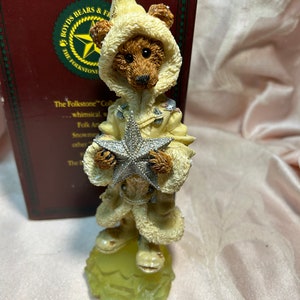 Puede incluir: Una figurita de oso blanco y marrón que lleva un abrigo blanco y sostiene una estrella plateada. El oso está de pie sobre una base verde. La colección Folkstone.