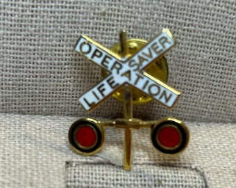 May include: A gold-colored metal pin with a white cross and the words "OPER SAVER LIFE ATION". The pin features two red circles with black borders and a vertical gold bar.