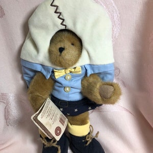 Op de afbeelding: Een bruine teddybeer die een blauw shirt draagt met een gele strik, een blauwe rok met witte sterren, gele sokken en blauwe schoenen. De beer draagt een witte, eivormige hoed met een bruin zigzagsteekdetail. De beer houdt een label vast met de tekst "The Original Teddy Bear Company".