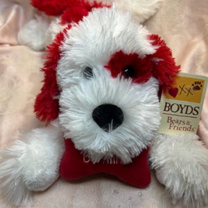 May include: A plush dog toy with white fur and red accents on the ears, around one eye, and on the bow tie. The toy has a black nose and the tag reads "BOYDS Bears & Friends".