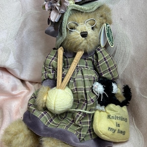 Puede incluir: Un oso de peluche vestido con un vestido a cuadros verdes y un gorro a juego, con gafas redondas pequeñas. El oso sostiene una bola de lana y una bolsa amarilla con un gato blanco y negro, que dice "Knitting is my bag".