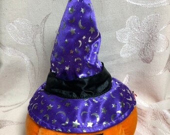 Puede incluir: Una calabaza de peluche naranja con una cara de calabaza negra que lleva un sombrero de bruja morado con estrellas y lunas plateadas.