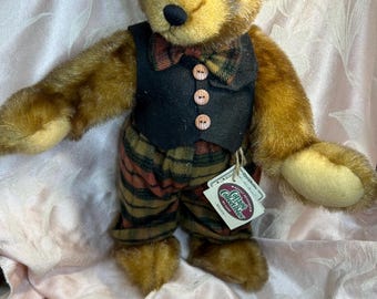 Puede incluir: Un oso de peluche marrón, con un chaleco negro con tres botones de madera, una pajarita a cuadros y pantalones a cuadros. El oso tiene una nariz negra y ojos pequeños y oscuros. Una etiqueta está adjunta al oso.