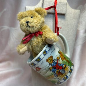 Puede incluir: Un pequeño oso de peluche con una cinta roja alrededor del cuello está metido dentro de una taza de cerámica decorativa. La taza presenta un diseño de oso de dibujos animados y un borde azul y blanco. Una cinta roja está unida en la parte superior para colgar.