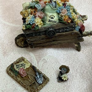 以下が含まれることがあります： カラフルな花、小さなテディベア、足跡のマークとBoydsのラベルが付いた袋で満たされた装飾用の樹脂製手押し車。その横には、シャベル、ピンクのバラ、Boyds Garden Stuffと書かれた看板、小さなクマの置物が置かれた編み込みマットがあります。