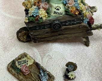 May include: A decorative resin wheelbarrow filled with colorful flowers, a small teddy bear, and a bag labeled Boyds with a paw print. Beside it lies a rectangular woven mat with a shovel, a pink rose, and a sign reading Boyds Garden Stuff, plus a small bear figurine.