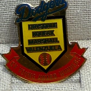 May include: A vintage Dodgers pin featuring the team's logo in blue and gold. The pin includes the names Drysdale, Koufax, Marshall, and Valenzuela, with a baseball graphic and the text "Cy Young Award Winners" in red.