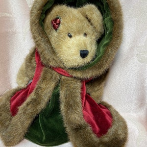 May include: A brown plush teddy bear wearing a green velvet hooded coat with a brown fur trim and a red velvet scarf. The bear has a red bow on its ear.