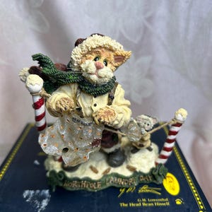 May include: A whimsical figurine of a cat dressed in winter attire, holding a laundry bag and pulling a small mouse. The cat wears a hat, scarf, and suspenders. The base has text that reads "North Pole Laundry Day".