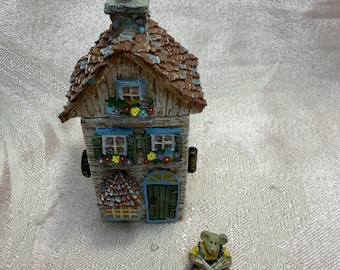 May include: A miniature house figurine with a brown shingled roof, blue window frames, and a green door. A small bear figurine is sitting and reading a book. The house has a chimney and decorative flower boxes.